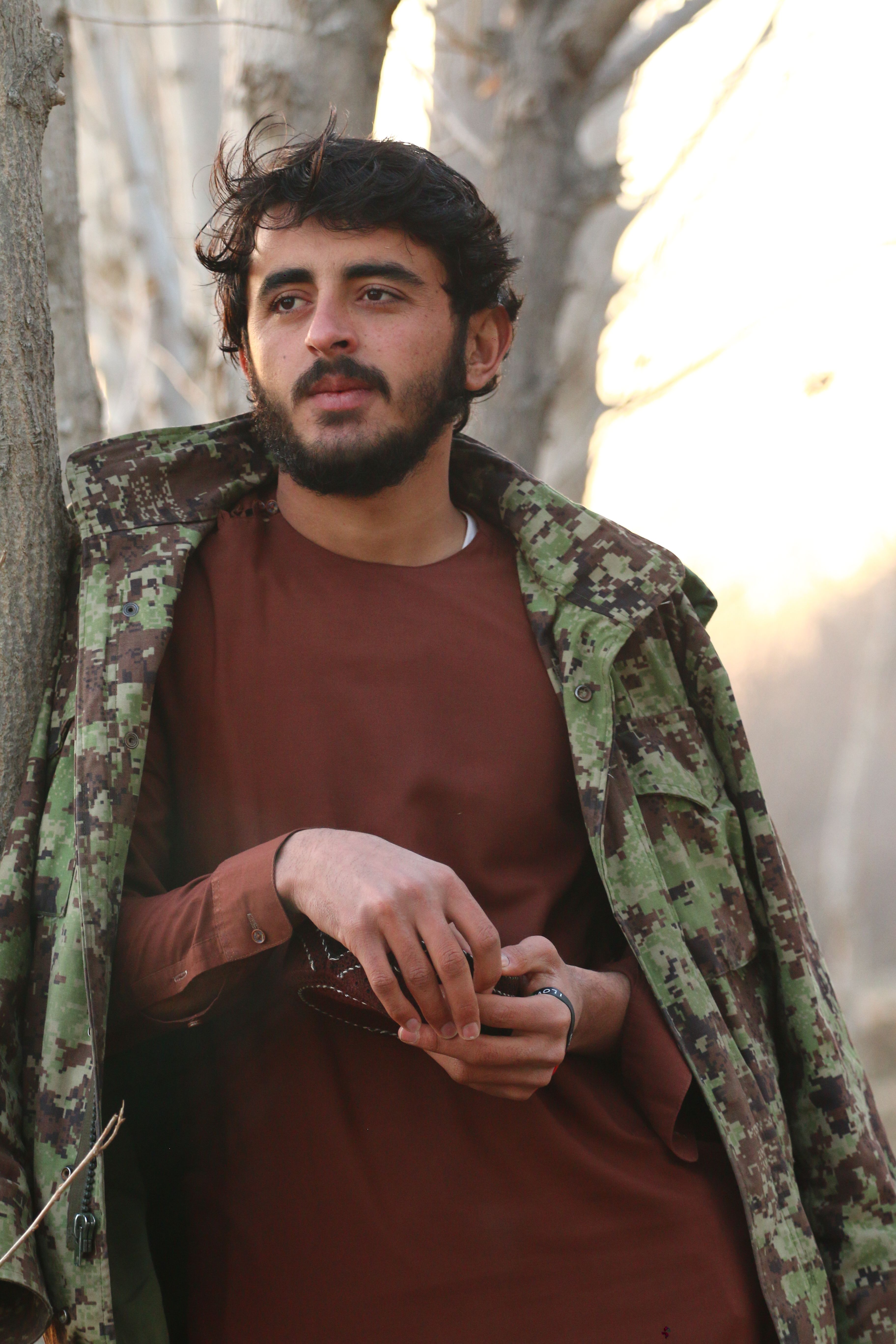Bearded man in camouflage jacket leans against tree during daytime outdoors.