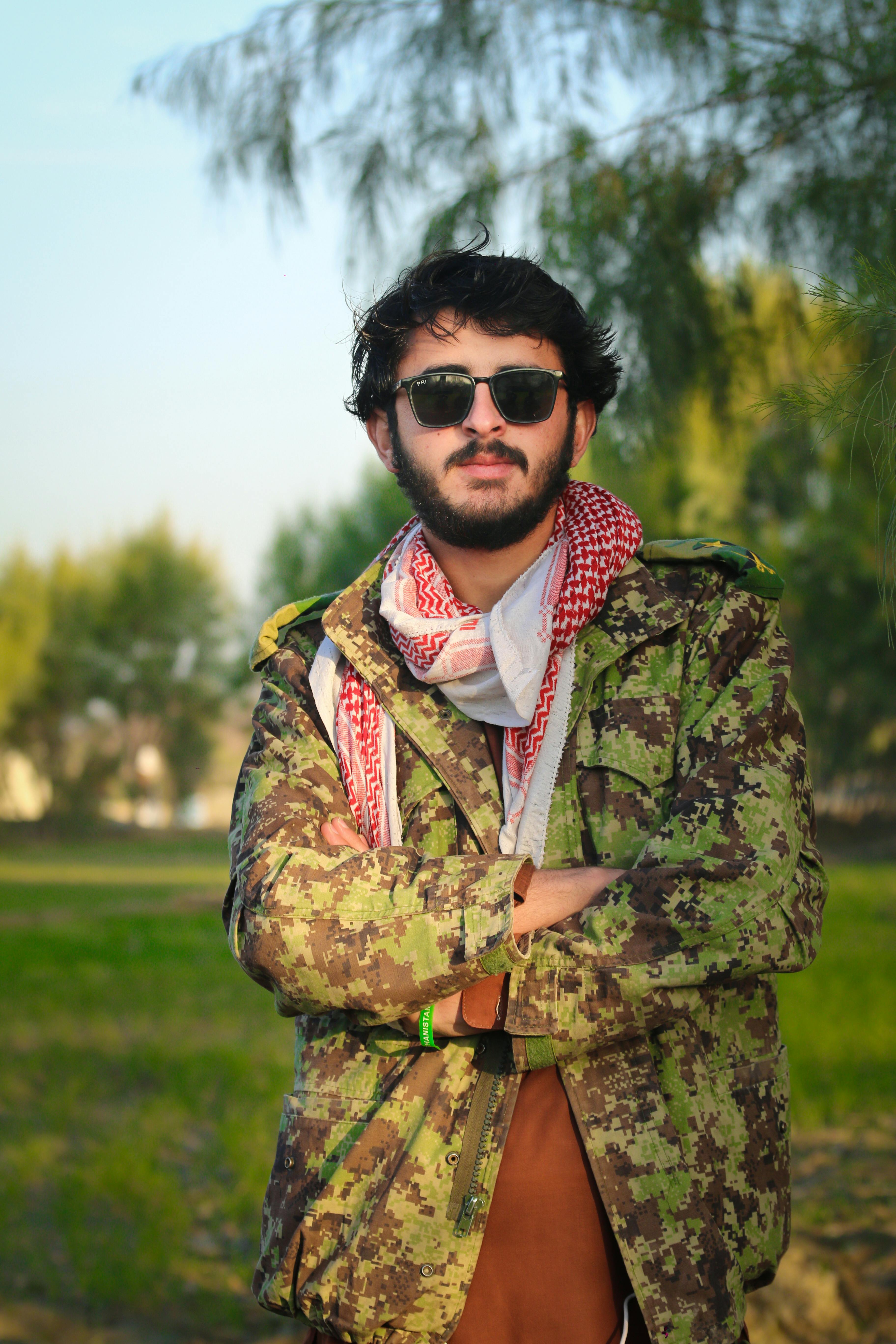 Man wearing camouflage jacket and sunglasses stands confidently outdoors in a sunny setting.