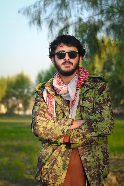 Free Man wearing camouflage jacket and sunglasses stands confidently outdoors in a sunny setting. Stock Photo