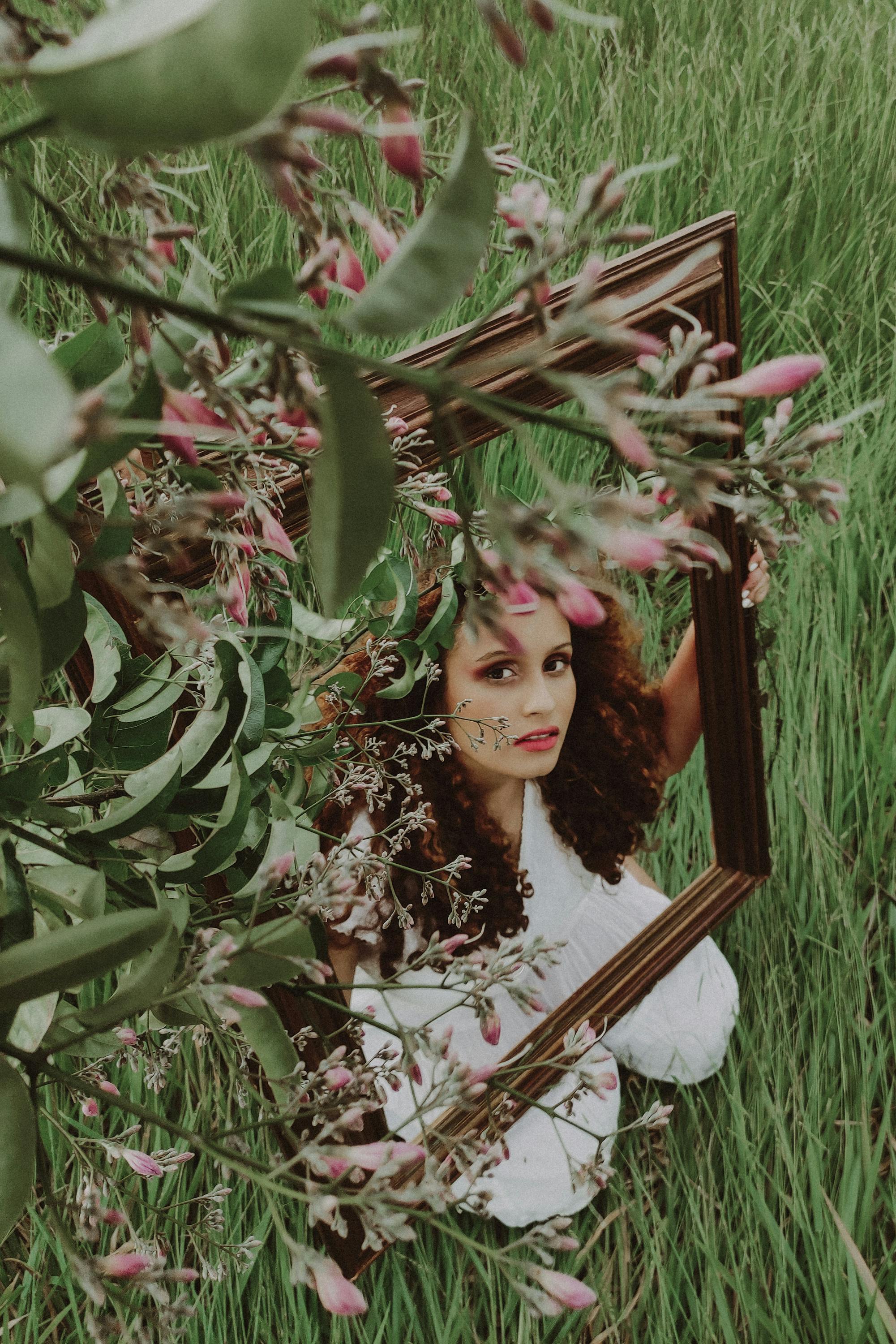 Artistic portrait of a woman holding a frame in a lush field, enveloped by flowers and leaves.