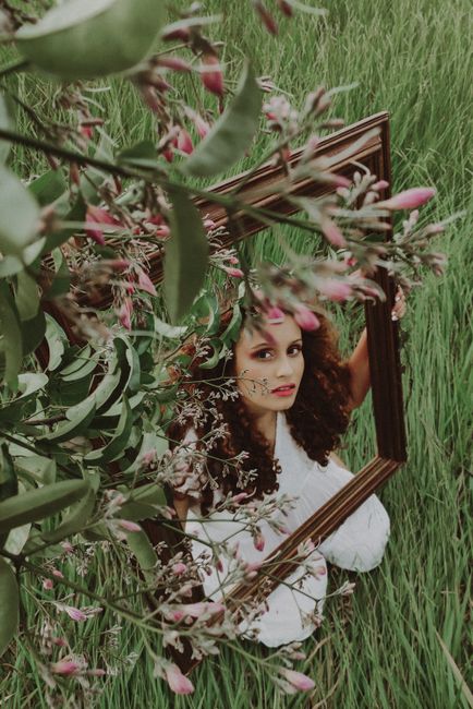 Artistic portrait of a woman holding a frame in a lush field, enveloped by flowers and leaves.