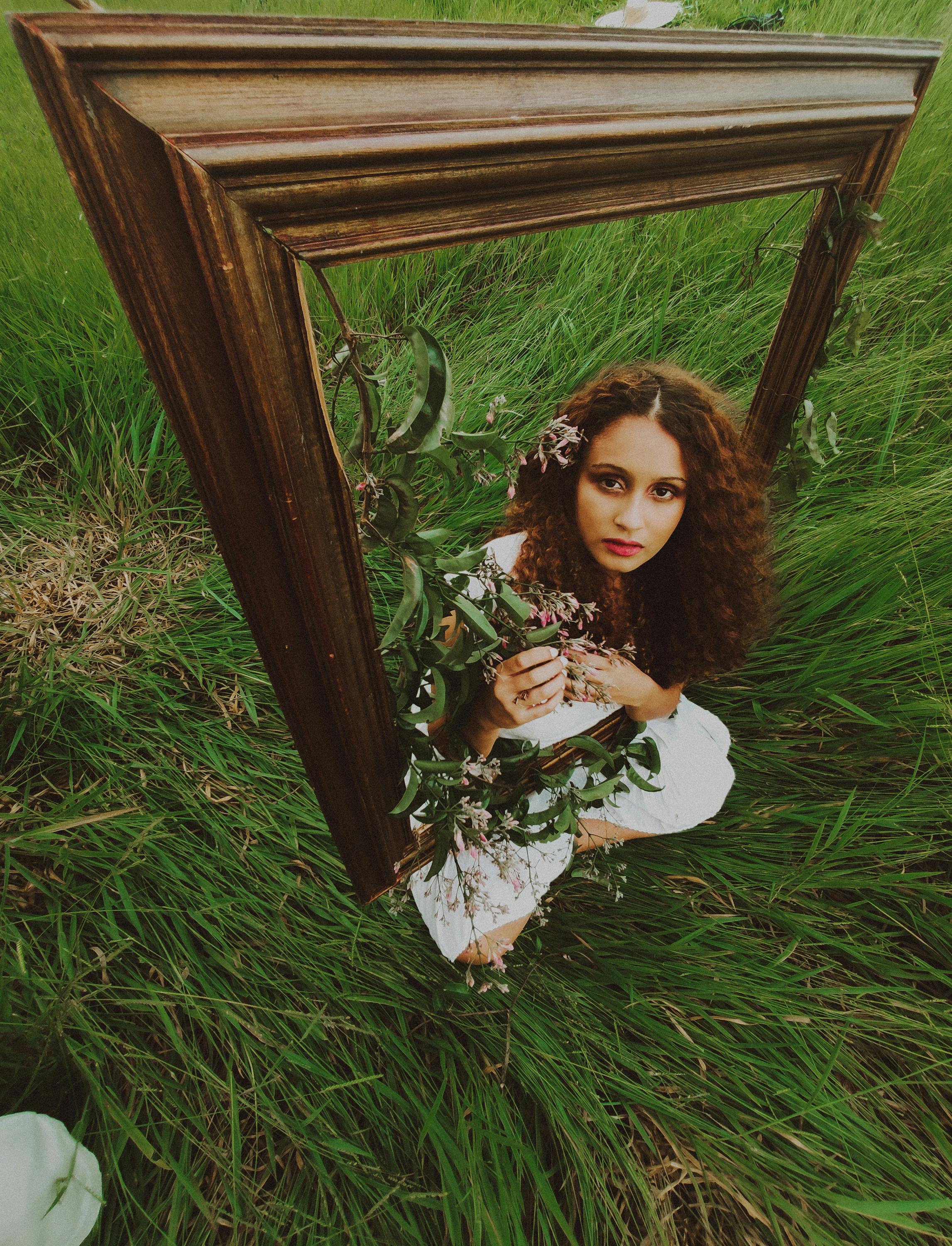 Artistic portrait of a woman with curly hair holding flowers, framed by a vintage wooden frame, set in a grassy field.