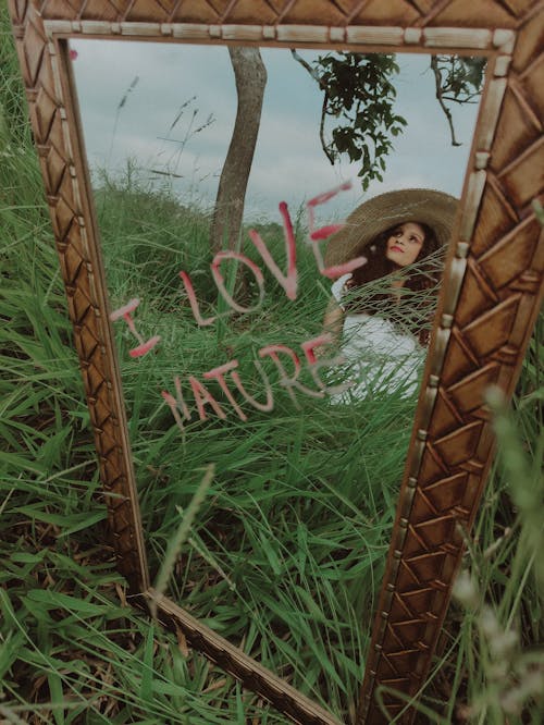Mirror Reflection of a Woman in White Dress and Straw Hat on Green Grass