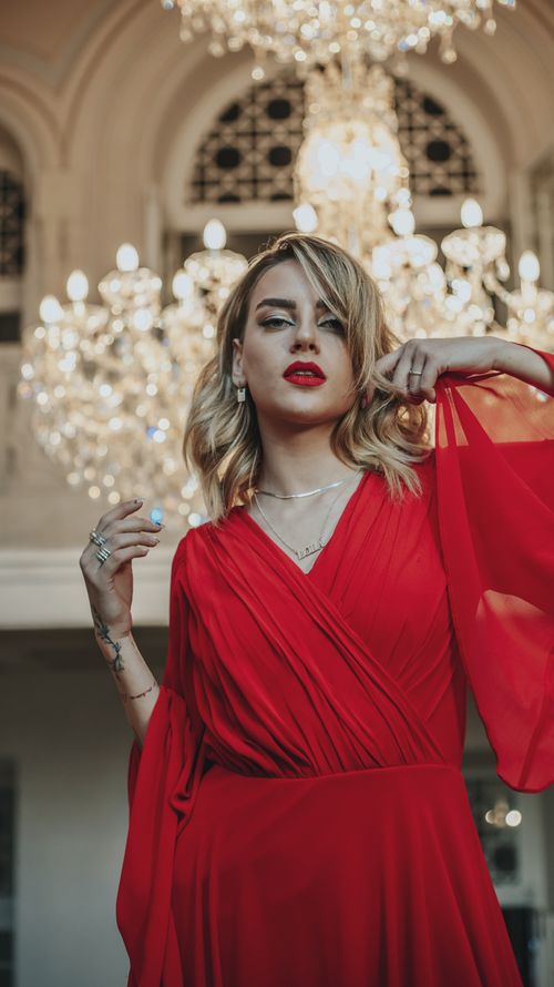 Free Stylish woman in red dress posing gracefully indoors with a chandelier backdrop. Stock Photo