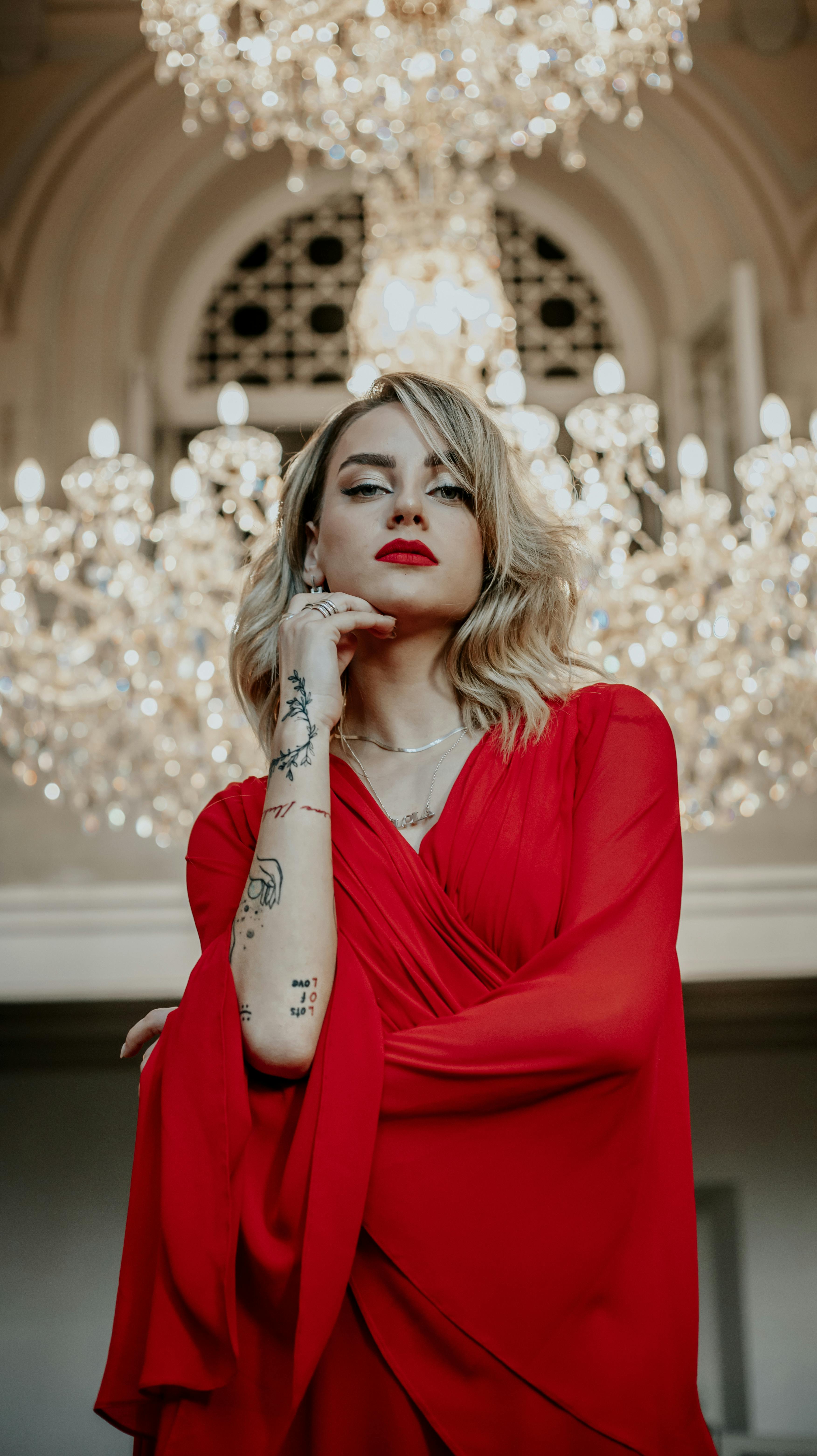 Portrait of a confident woman in a stylish red dress with tattoos, standing elegantly under a chandelier.