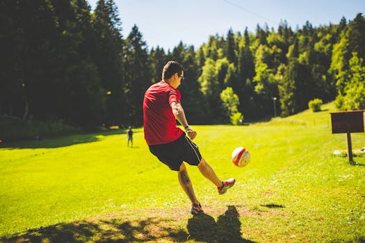 Man in a red shirt kicking soccer ball on sunny day in a park, surrounded by lush trees.