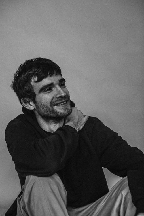 Free Black and white portrait of a young, bearded man smiling and sitting indoors with his hand on his shoulder. Stock Photo