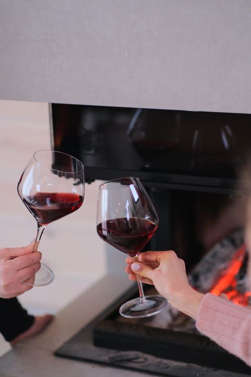 Free Two people toasting with red wine glasses by a warm fireplace, creating a cozy atmosphere. Stock Photo