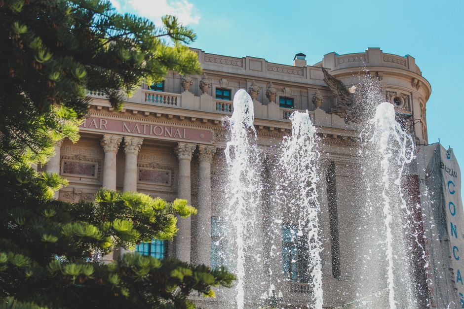Una gran fachada arquitectónica con una fuente que fluye y exuberantes árboles verdes bajo un brillante cielo azul.