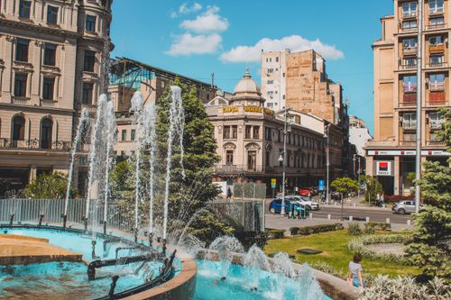 Free Sunny day in an urban plaza with a fountain, classic architecture, and bustling streets. Stock Photo
