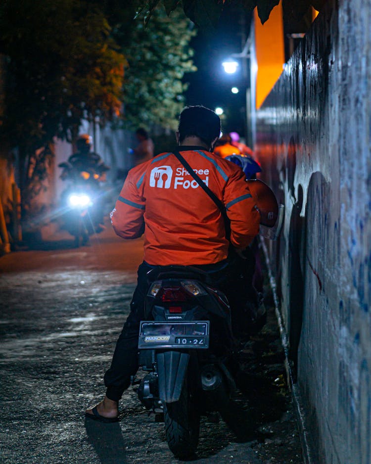 A Back View Of A Man In Orange Jacket Riding A Motorcycle On The Street At Night