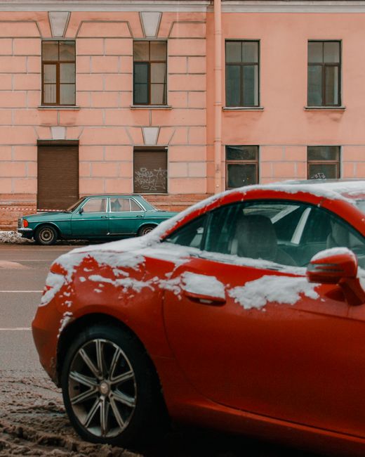 A striking red sports car with snow cover parked on a city street in winter, contrasting a vintage building.