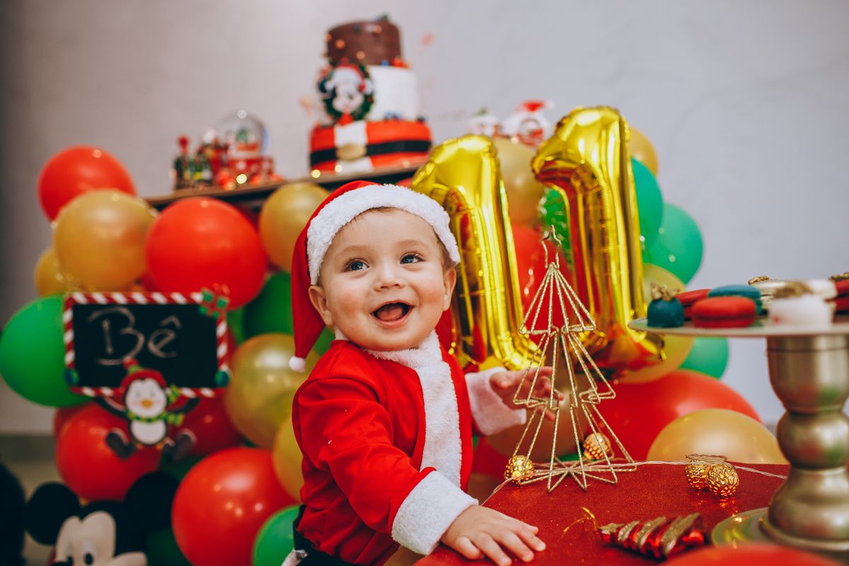 Adorable baby wearing Santa costume enjoying a colorful festive setting with balloons and Christmas decorations. Stock Photo.