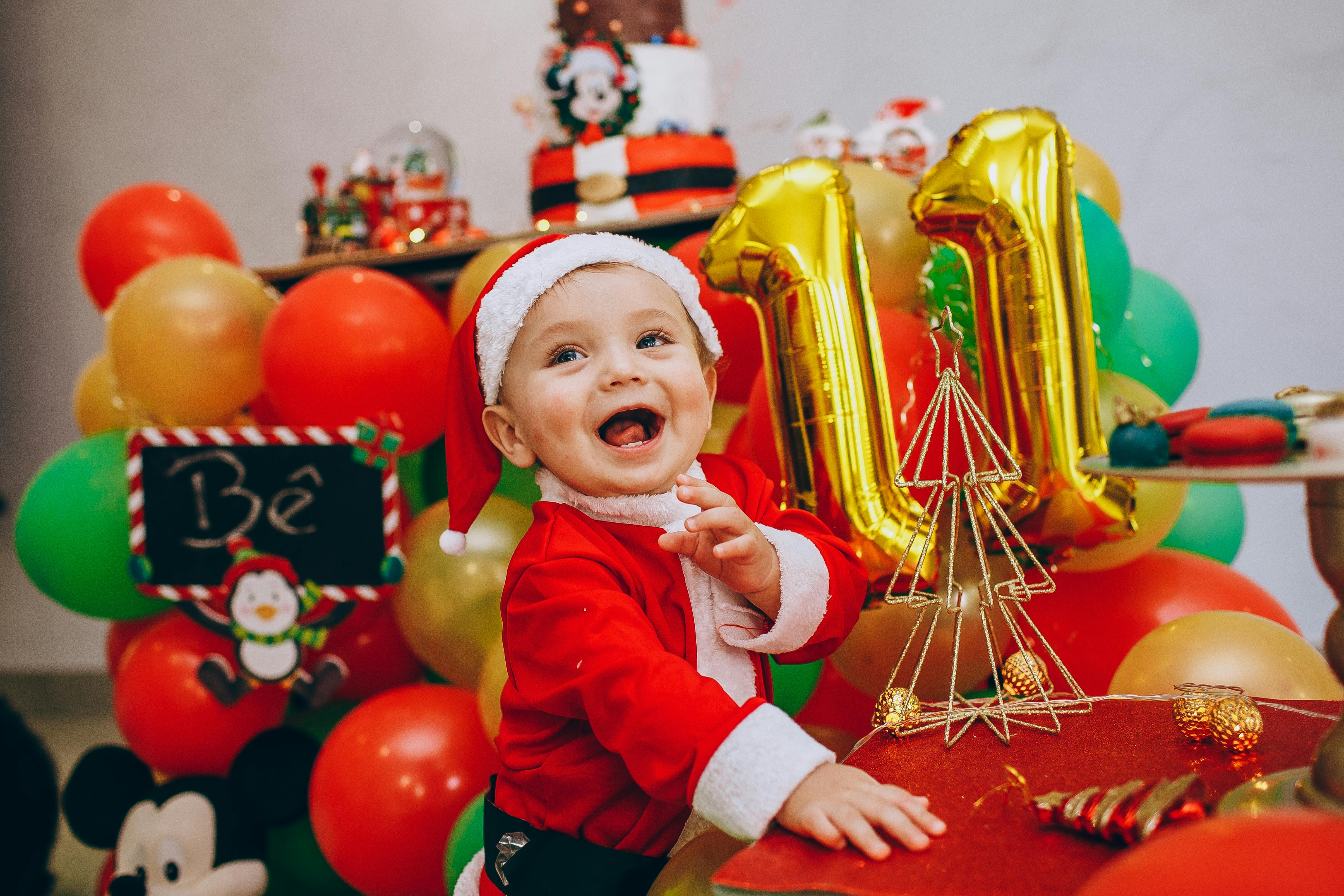 Joyful baby dressed as Santa Claus amidst colorful Christmas decorations indoors.