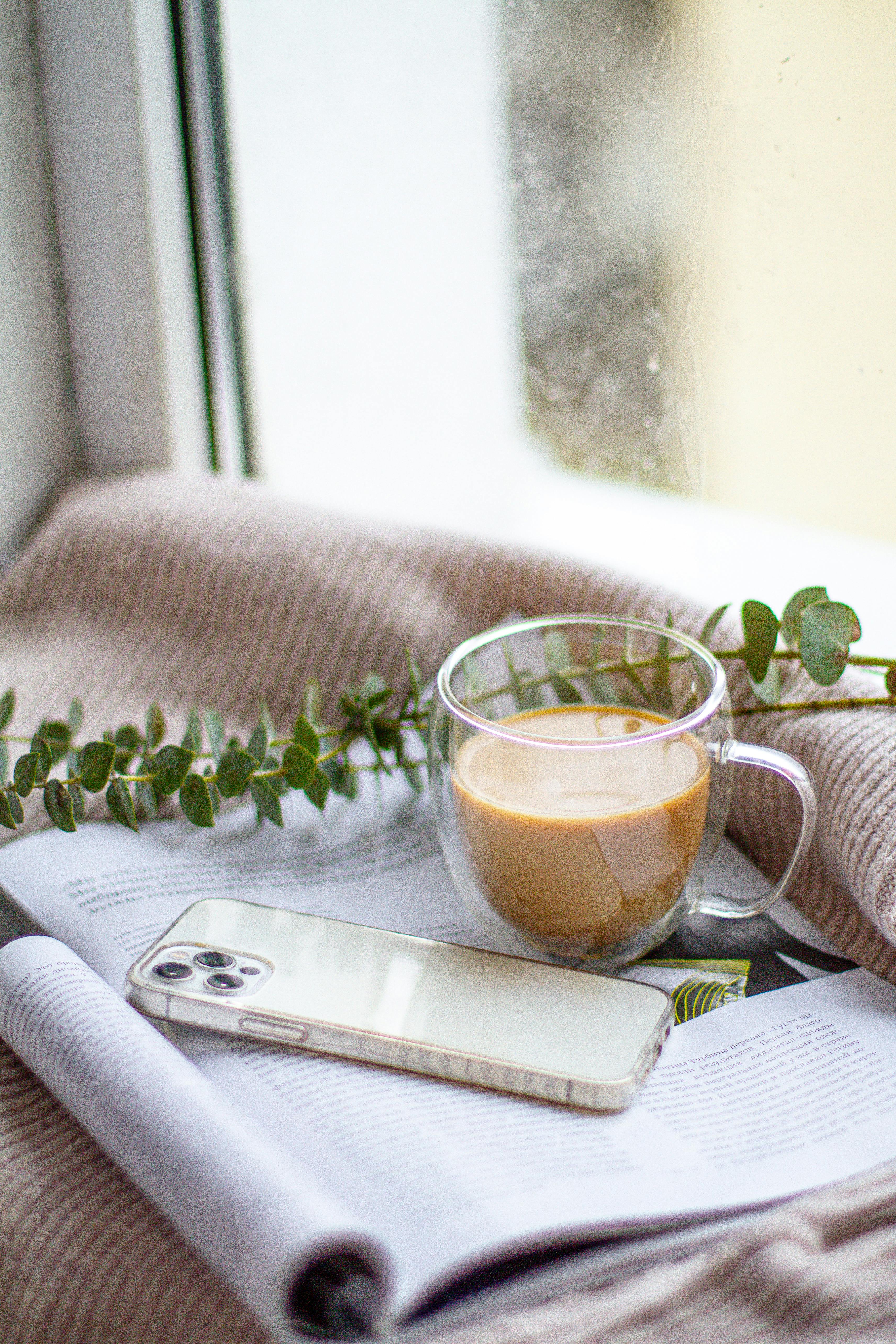 a clear glass cup beside a smartphone on a magazine by the window