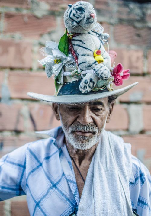 Free Portrait of an elderly man wearing a hat decorated with a stuffed tiger and flowers, set against a brick wall. Stock Photo
