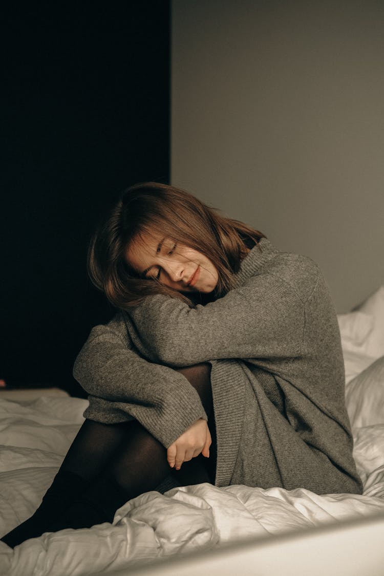 Woman Sitting On Bed With Head On Bent Knees And Smiling