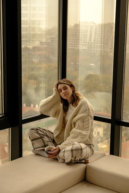 Free Woman Wearing Big Cardigan and Checkered Pants Sitting Crossed Legged by Office Window Stock Photo