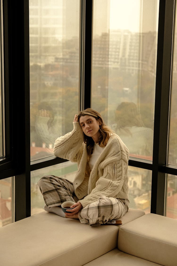 Woman Wearing Big Cardigan And Checkered Pants Sitting Crossed Legged By Office Window