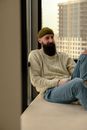 Bearded Man in Hat Sitting Relaxed on Sofa by Window in Office Room and Smiling