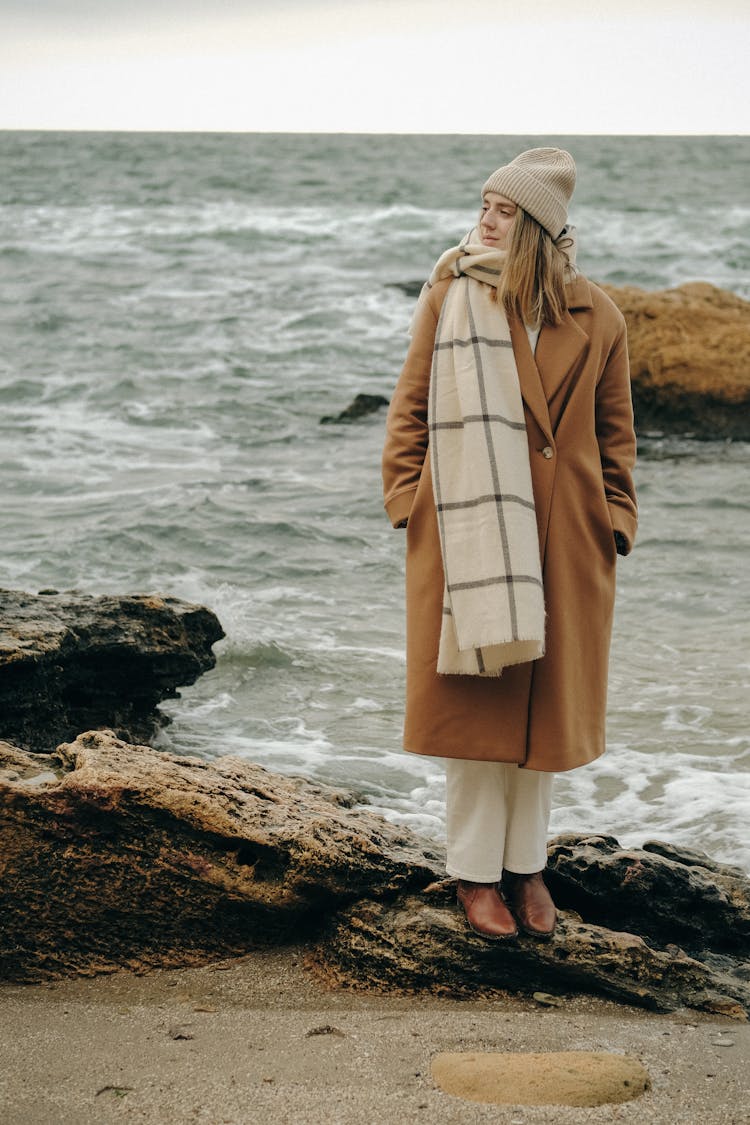 Blond Woman With Long Straight Hair In Knitted Hat And Warm Clothes Standing On Beach At Sea