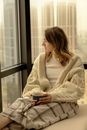 Blond Woman in Oversized Cardigan Sitting Relaxed on Sofa with Cup of Tea in Hands 
