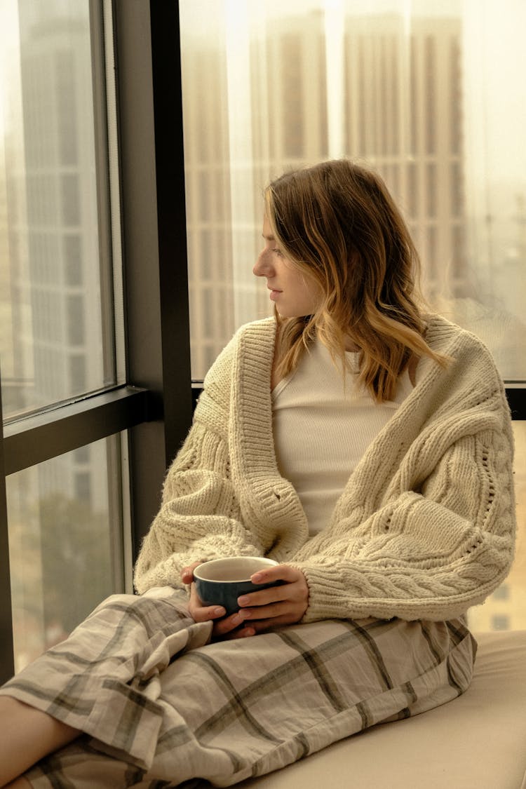 Blond Woman In Oversized Cardigan Sitting Relaxed On Sofa With Cup Of Tea In Hands 