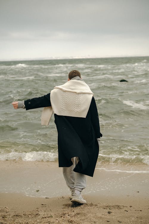 Unrecognizable Man Wearing Black Coat Standing on Beach with Stretched Arm to Side