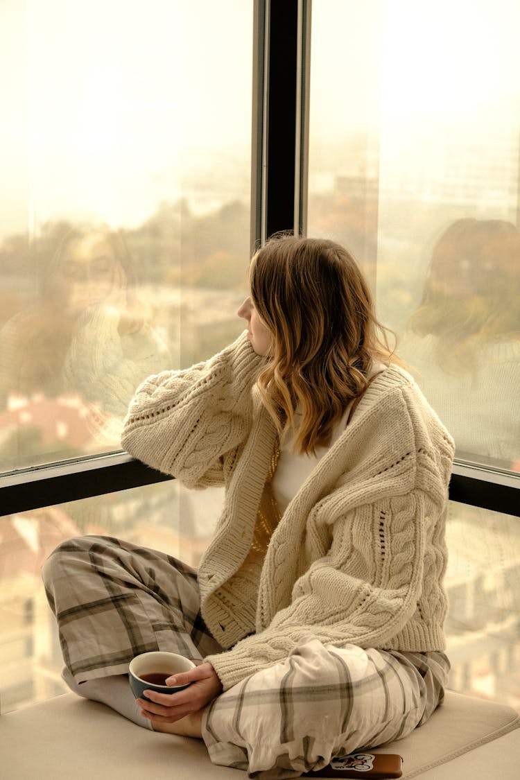 Cross Legged Sitting Woman Holding Cup Of Tea Looking Through Window