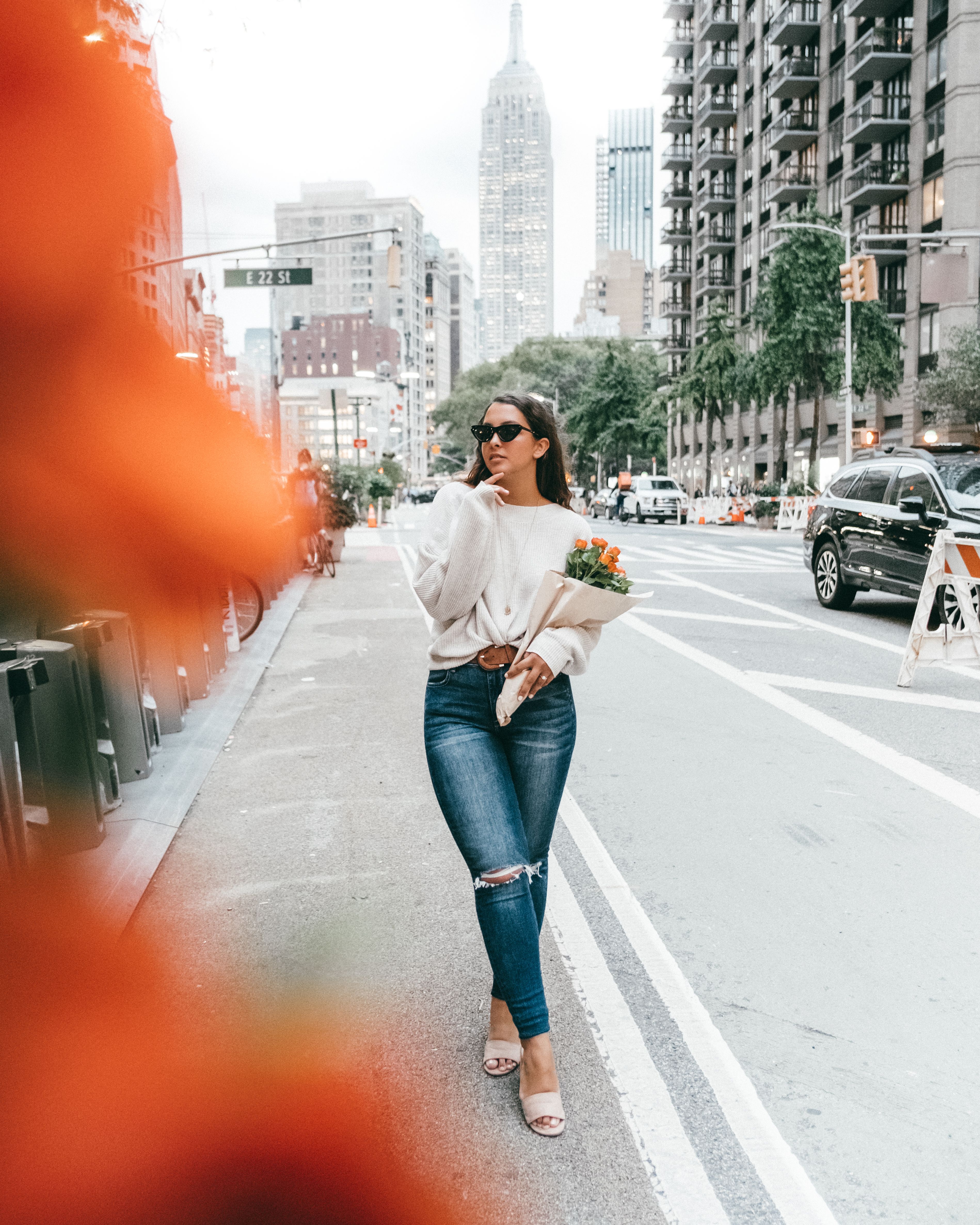 Chic woman in sunglasses with flowers strolling through a vibrant New York City street.