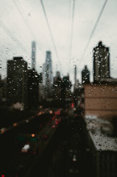 View of a blurred city skyline through raindrop-covered window, creating a moody urban atmosphere.