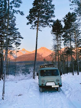 A vintage van parked on a snowy forest path against a mountain backdrop at sunrise.