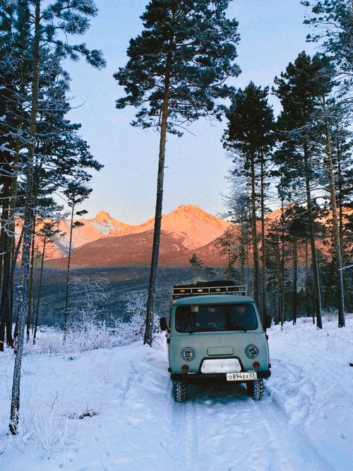 Free A vintage van parked on a snowy forest path against a mountain backdrop at sunrise. Stock Photo