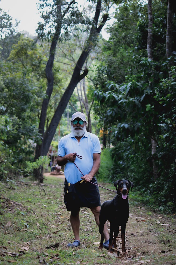 A Man In White T-shirt And Black Shorts Holding The Leash Of A Black Short Coated Dog