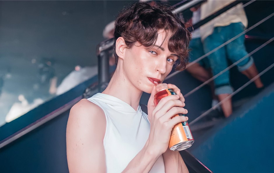 Person with short hairstyle drinking from a can indoors, wearing a sleeveless top.