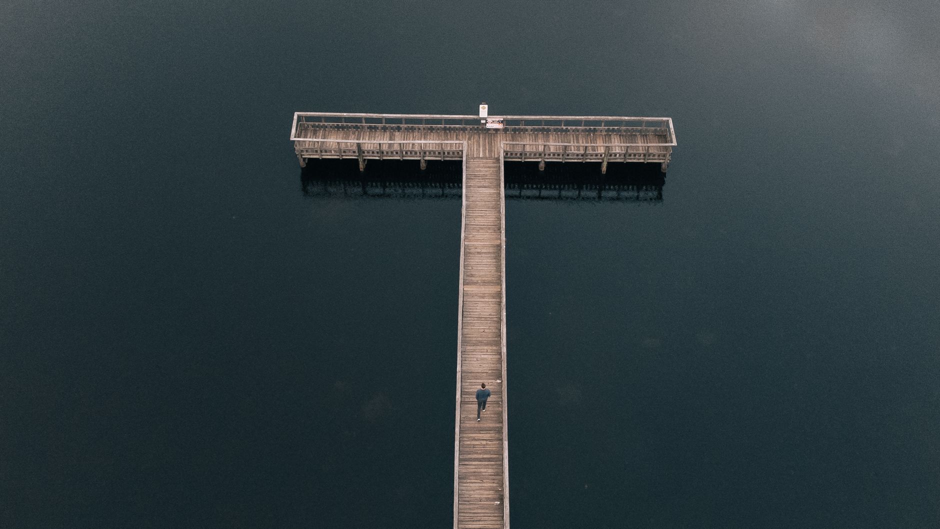 Serene aerial view of a wooden dock extending over a calm body of water with a lone figure walking.