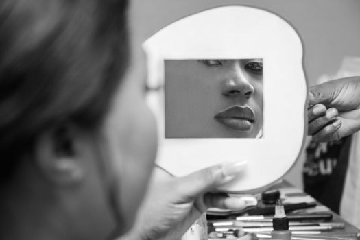 Black and white photo of a woman looking into a mirror, applying makeup.