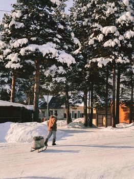 Person in winter clothing pulling a sleigh on snow-covered ground with snowy trees in the background.