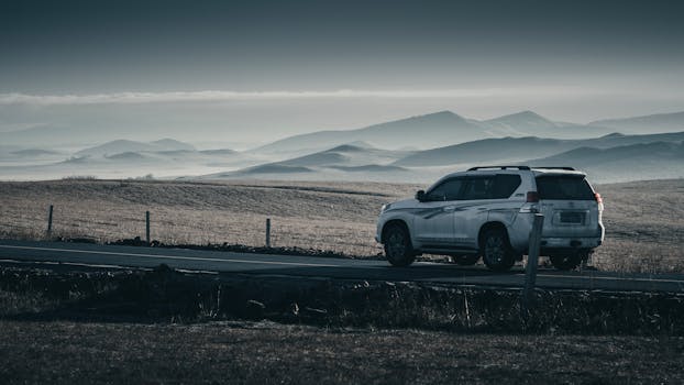 A white SUV parked on a scenic road with a backdrop of rolling hills and mountains under a cloudy sky.