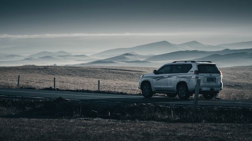 Free A white SUV parked on a scenic road with a backdrop of rolling hills and mountains under a cloudy sky. Stock Photo