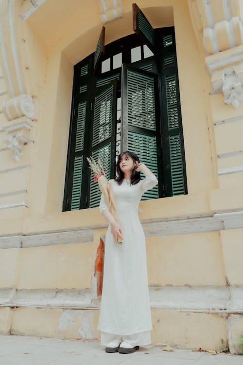 A Woman in White Dress Standing Beside a Building Window