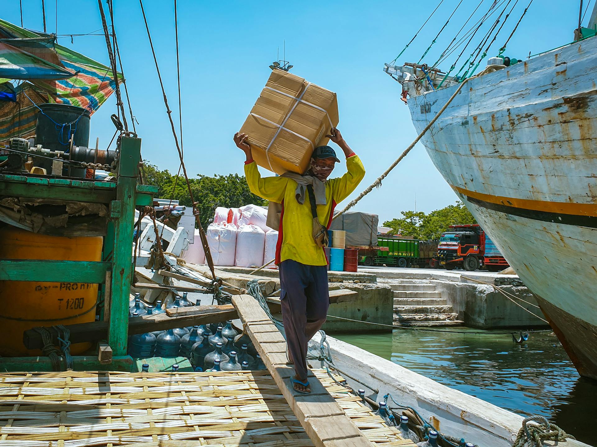 A dock worker balances a cardboard package on his head at a bustling Indonesian port, showcasing vibrant maritime activity.
