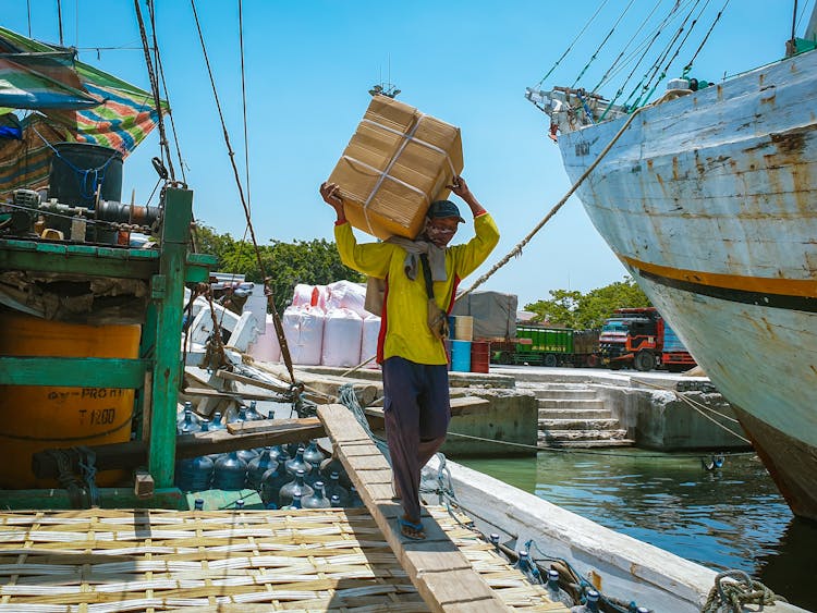 A Man Carrying A Cardboard Box Near A Boat