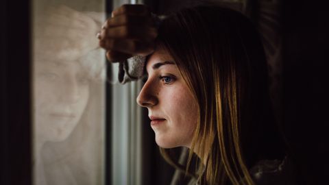 A contemplative young woman looks out a window with a reflective expression, indoors.