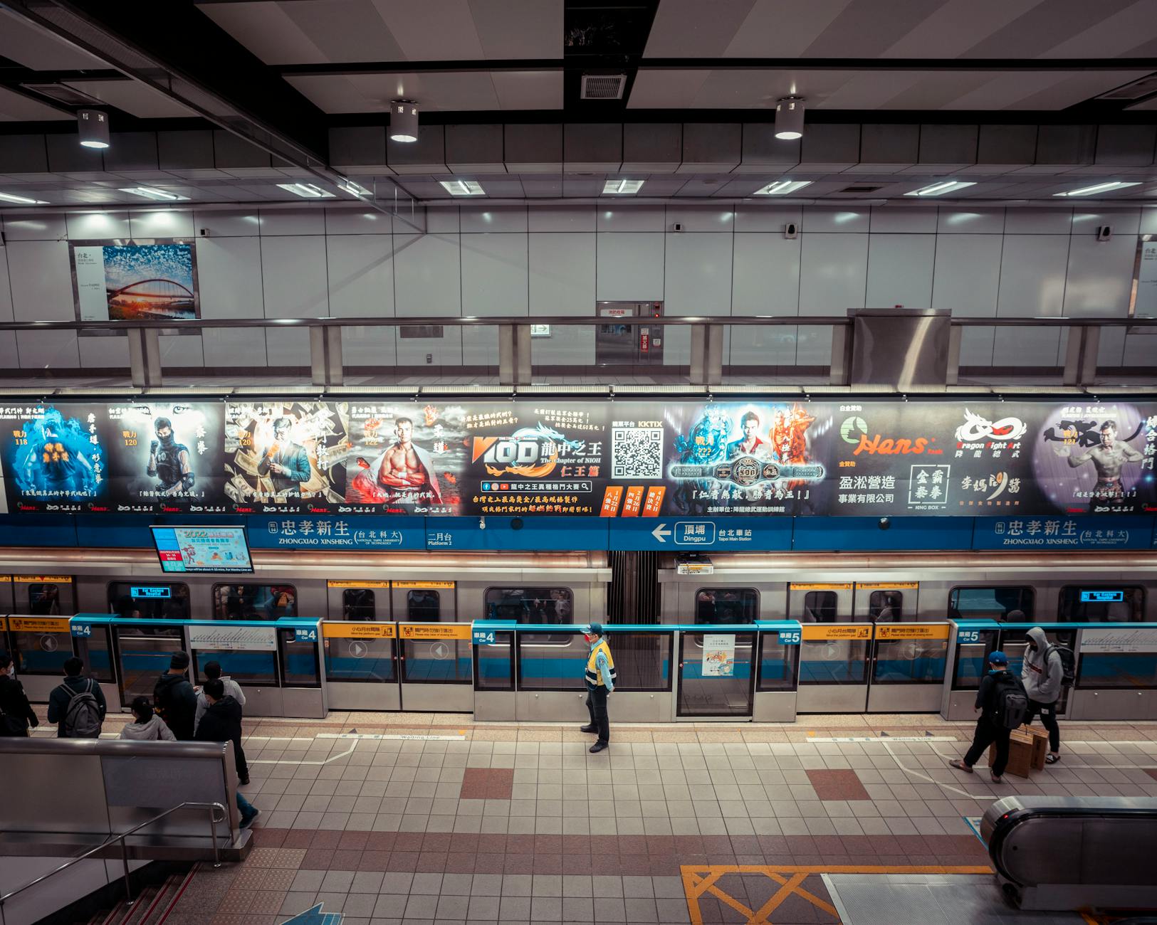 High angle view of a metro station with colorful advertisements and commuters standing and waiting.