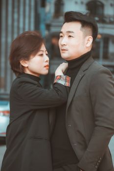 A fashionable couple poses in matching blazers and turtlenecks in an urban environment.