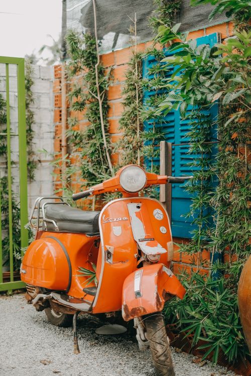 Free Retro orange scooter parked by a colorful wall with plants and blue shutters. Stock Photo