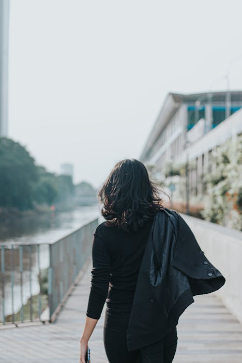 A Woman Holding Black Jacket Standing Near Body of Water