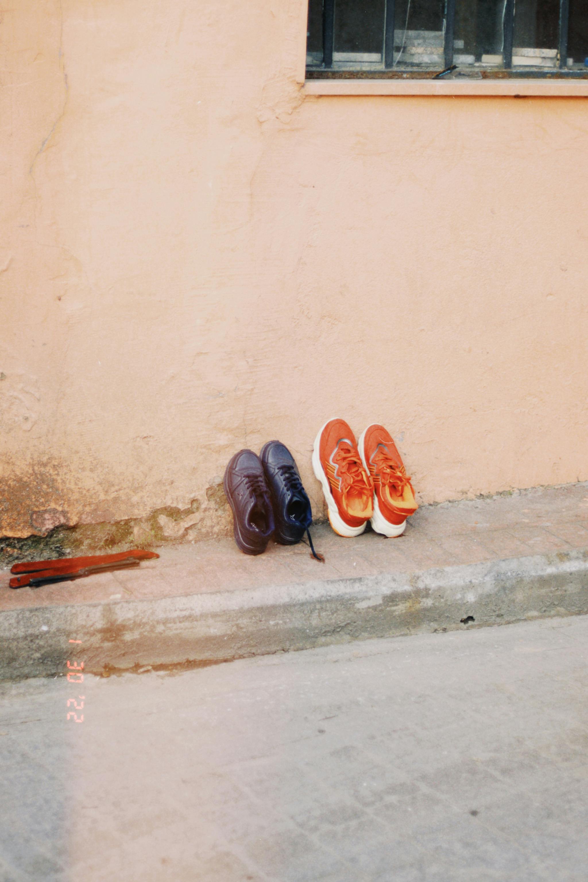 Two pairs of shoes leaning against a pastel wall on an urban street.