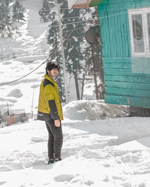 Free Adult man wearing a puffer jacket standing in a snowy landscape next to a green wooden house. Stock Photo